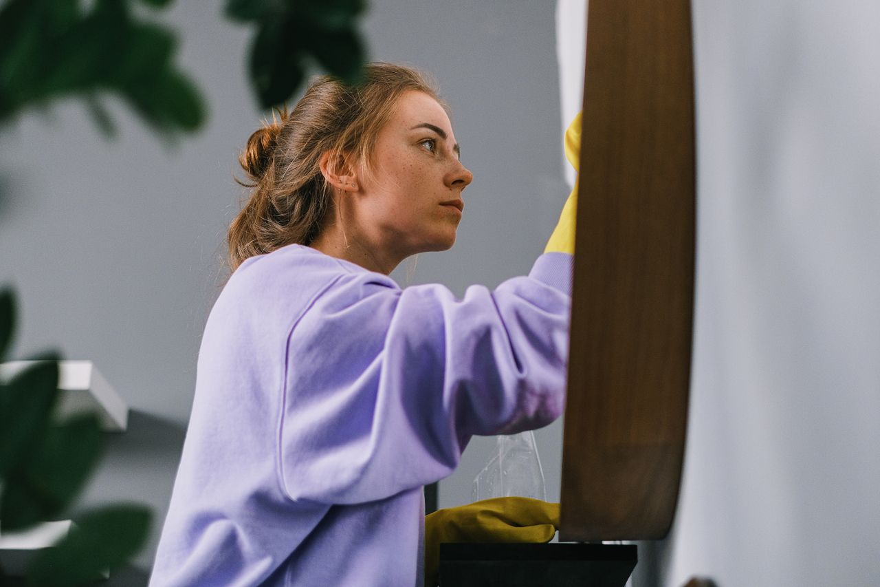 Side view of a young, focused woman in latex gloves standing in a bright room during the day and cleaning a mirror